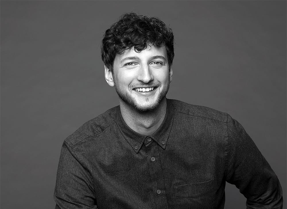 Porträtfoto von Dominik Keller – lächelnder Mann mit lockigem Haar in grauem Strickpullover