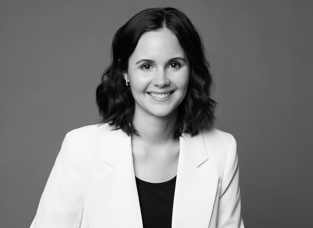 Porträtfoto von Olivia Zeitler – lächelnde Frau mit welligem, schulterlangemHaar in weißem Blazer