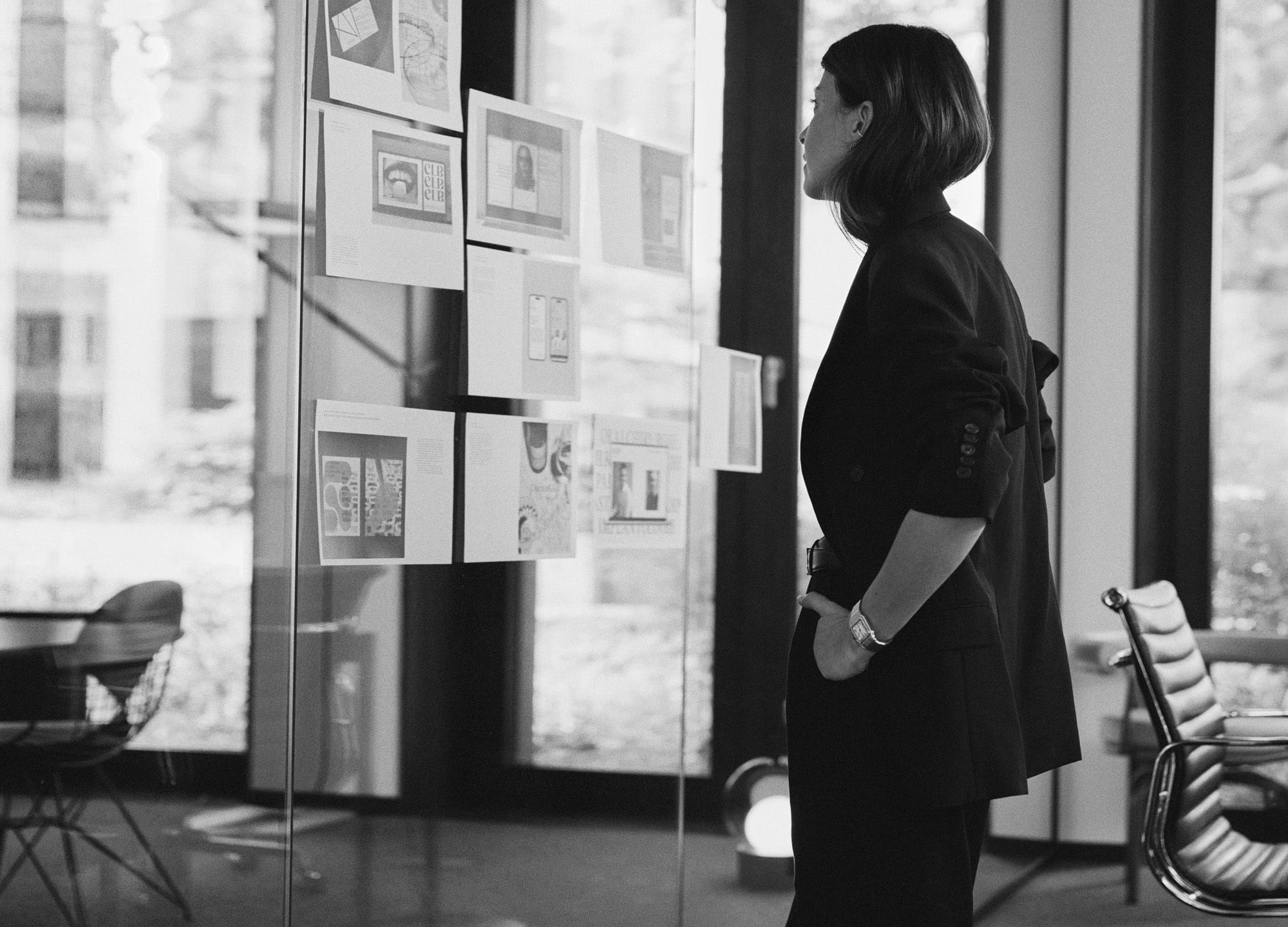 Frau in schwarzer Kleidung betrachtet konzentriert ein Moodboard an einer Glaswand in einem Büro – Schwarz-Weiß-Fotografie