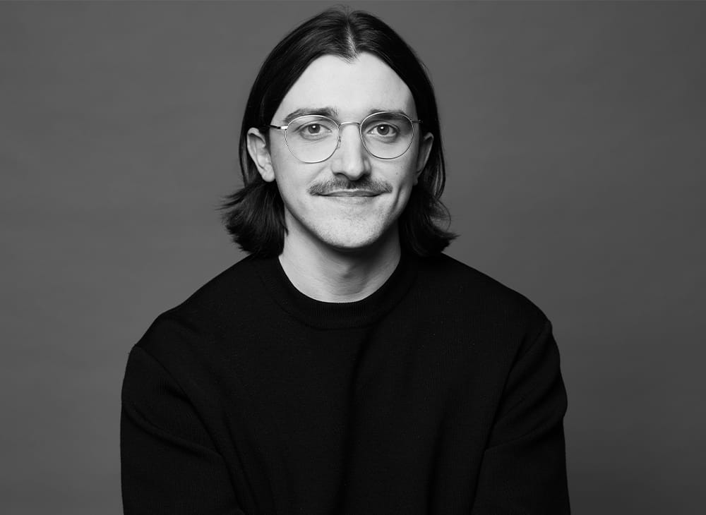 Porträtfoto von Thomas John – junger Mann mit langen Haaren, Bart und Brille in schwarzem Oberteil